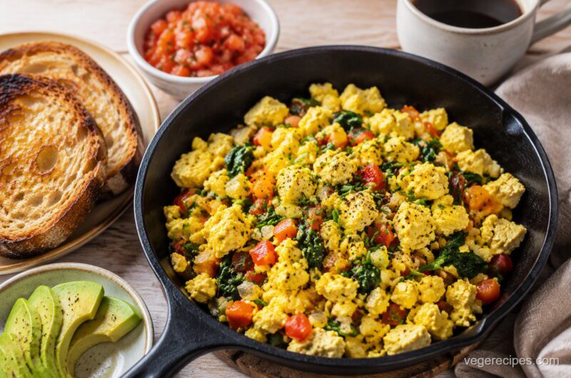 Golden tofu scramble with red bell pepper, spinach and chives in a cast-iron skillet, served with toast, avocado and salsa