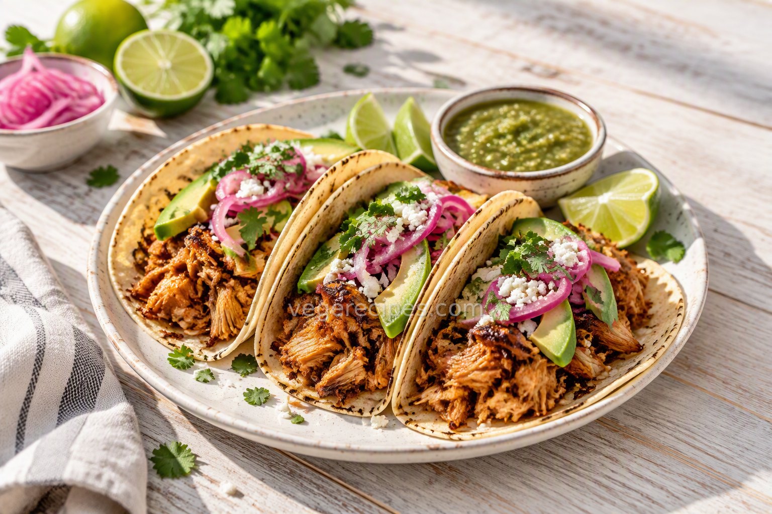 Three jackfruit carnitas tacos with avocado, pickled red onions, cilantro and cotija, served with lime wedges and salsa verde
