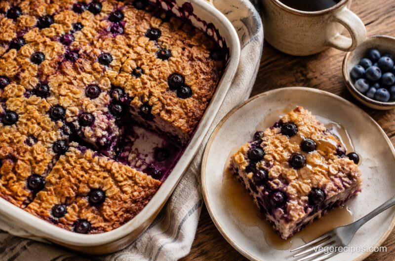 A baking dish of golden baked blueberry oatmeal with a slice served on a plate drizzled with maple syrup, coffee and fresh blueberries alongside