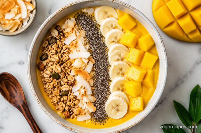 Overhead shot of a mango smoothie bowl topped with rows of granola, toasted coconut flakes, chia seeds, banana slices, and fresh mango cubes