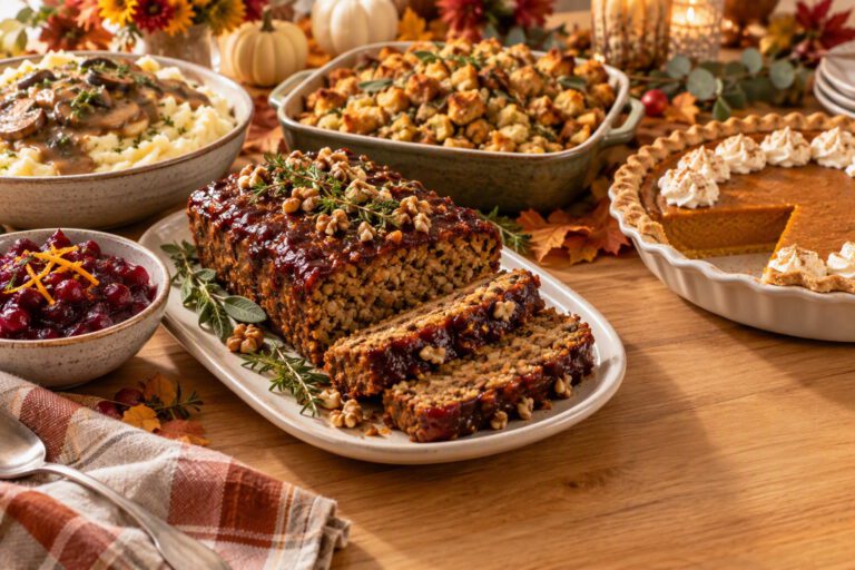 Vegetarian Thanksgiving table with a glazed lentil walnut loaf on a platter with two slices cut, surrounded by mashed potatoes with mushroom gravy, herb stuffing, cranberry-orange sauce and a pumpkin pie with one slice missing, with small pumpkins and autumn leaves